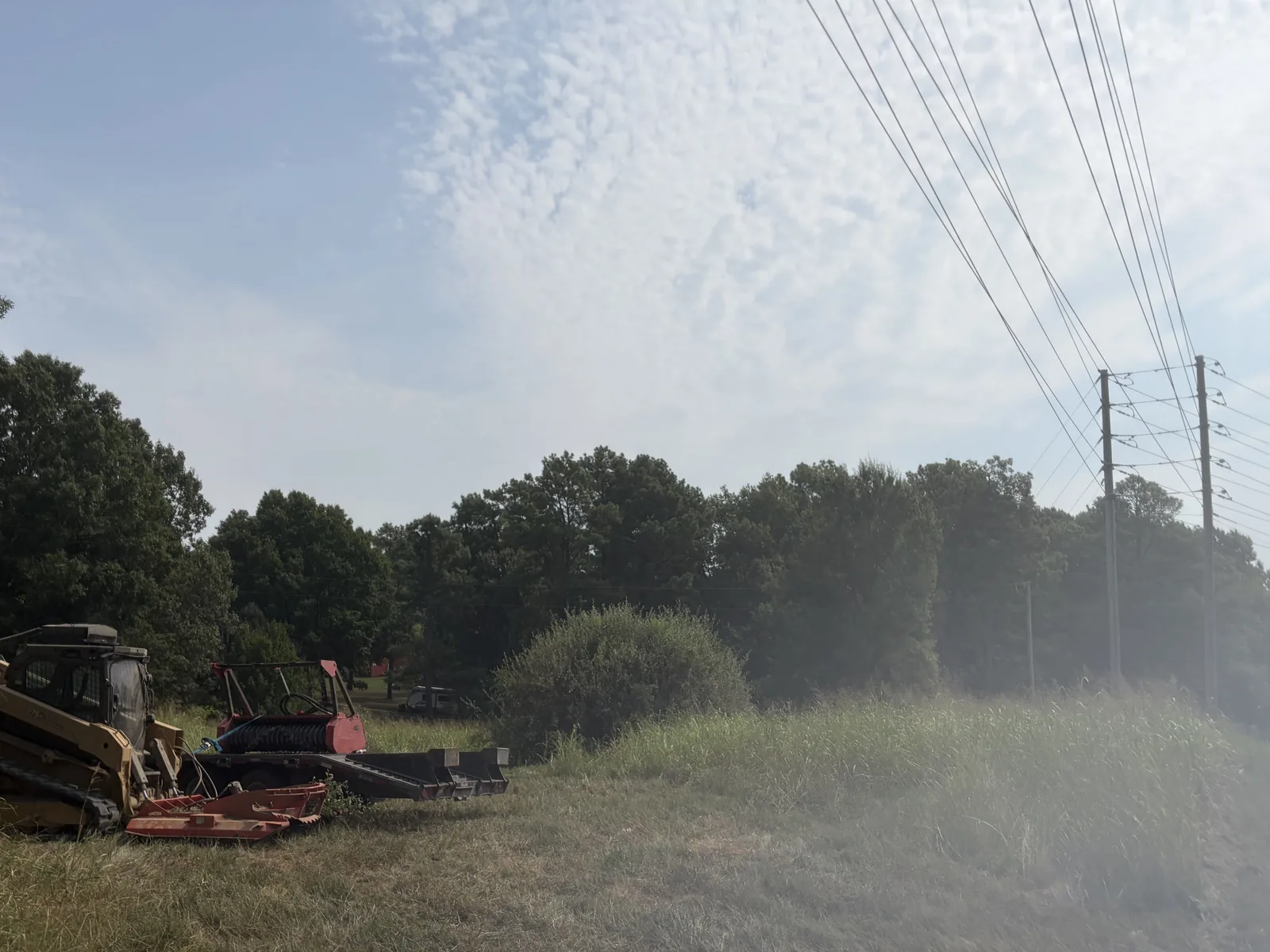 Compact track loader parked on freshly mowed grass with a brush cutter and a second attachment beside it in Northeast Arkansas