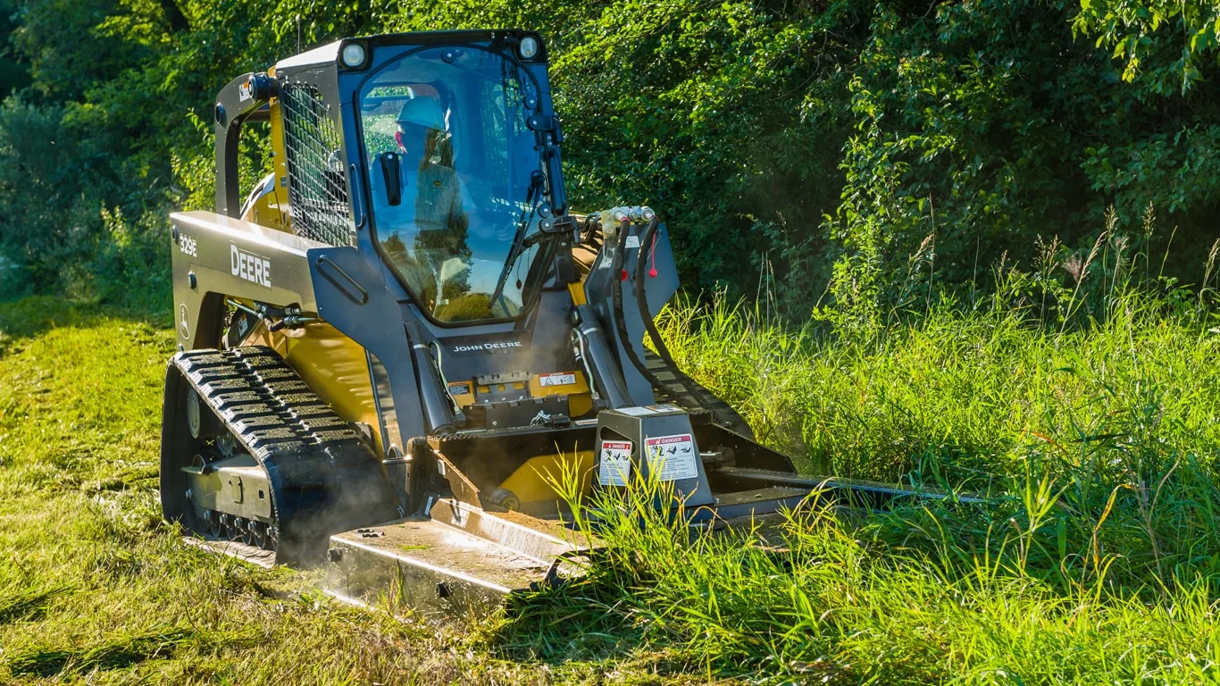 Compact track loader running a rotary brush cutter through tall grass on a grassy bank