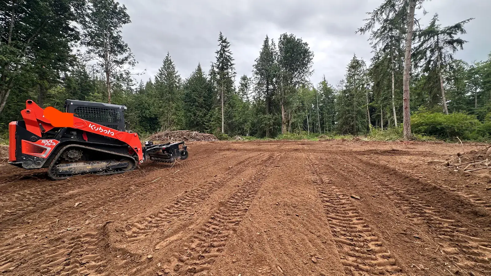 Freshly graded dirt lot combed smooth at the edge of the timber with a track loader parked to the side