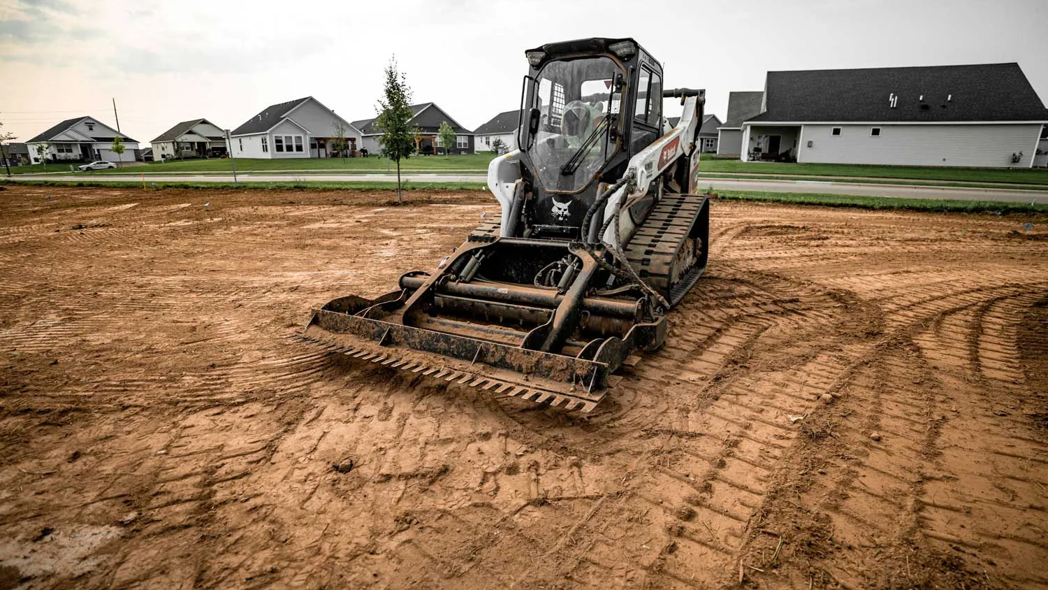Compact track loader pulling a landscape rake across a graded dirt yard