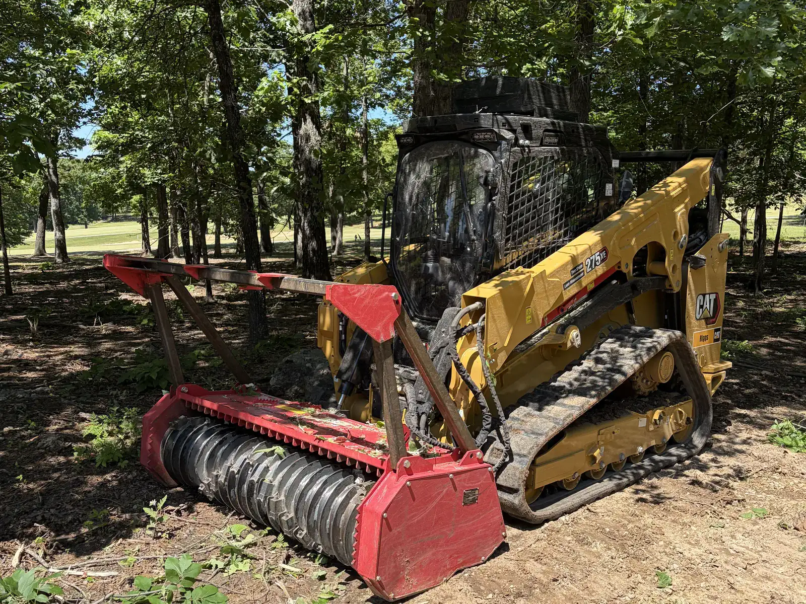 Compact track loader fitted with a red drum mulcher head parked on bare dirt in the woods in Northeast Arkansas