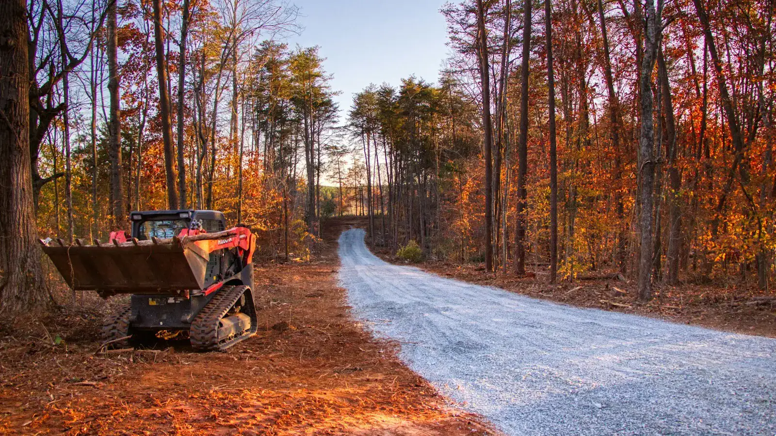 New gravel lane cut through fall timber with a compact track loader parked on the cleared shoulder