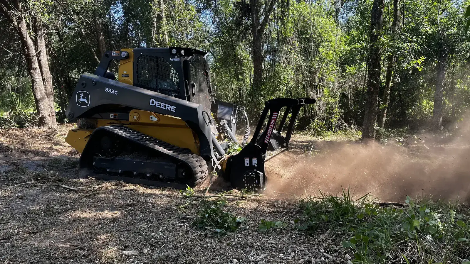 Compact track loader mulching through undergrowth in open timber, kicking up a cloud of dust